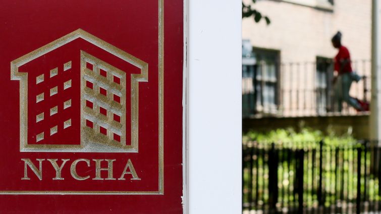 A NYCHA sign in front of a patch of grass with a person walking into a housing development in the background.