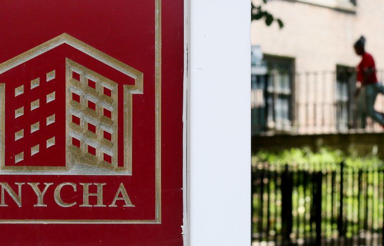 A NYCHA sign in front of a patch of grass with a person walking into a housing development in the background.