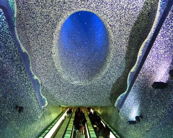 An escalator with a blue and purple tile mosaic on the ceiling overhead.