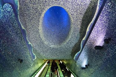 An escalator with a blue and purple tile mosaic on the ceiling overhead.