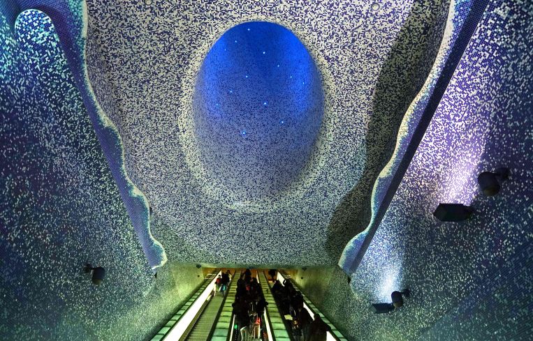 An escalator with a blue and purple tile mosaic on the ceiling overhead.