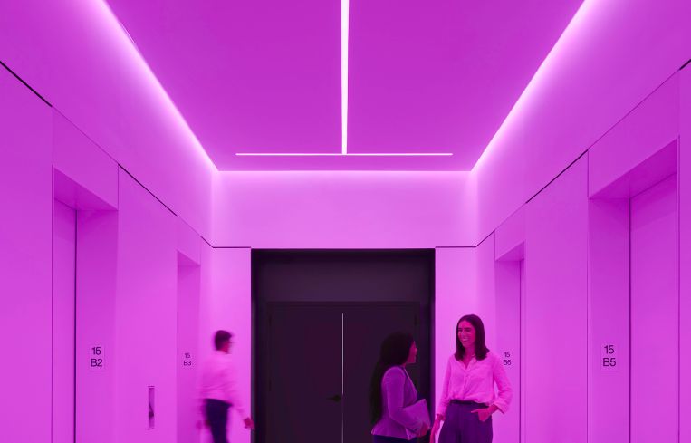 Three people stand next to a bank of elevators cast in a pink glow.