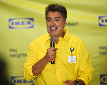 A man in a yellow button-up shirt speaking into a microphone in front of an Ikea-branded backsplash.