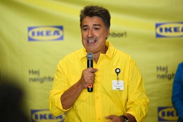 A man in a yellow button-up shirt speaking into a microphone in front of an Ikea-branded backsplash.
