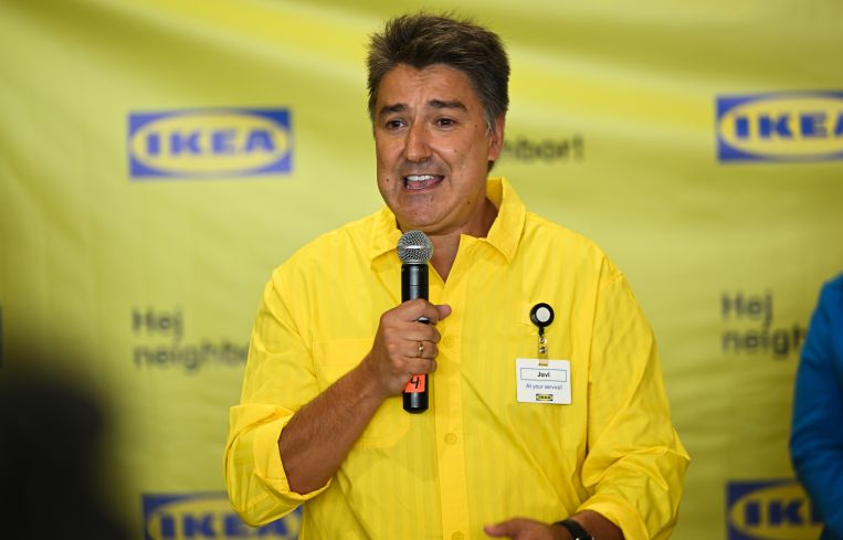 A man in a yellow button-up shirt speaking into a microphone in front of an Ikea-branded backsplash.