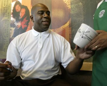 A man sitting down, taking a coffee mug from someone, and preparing to sign it.