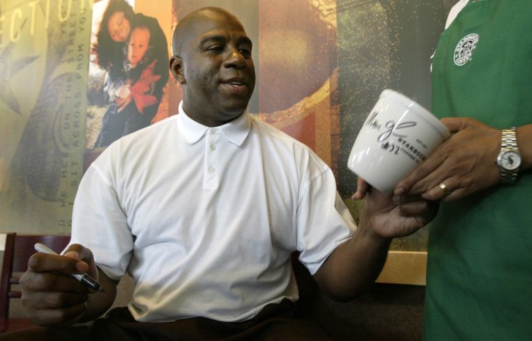 A man sitting down, taking a coffee mug from someone, and preparing to sign it.