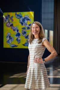 A woman standing and smiling, next to a painting.