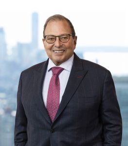 A smiling man in a suit in front of a window with buildings behind him.