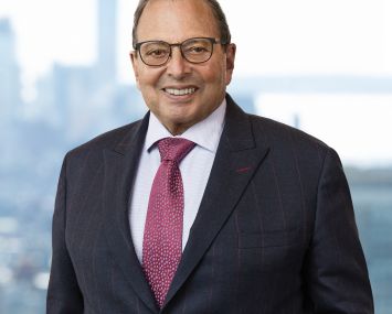 A smiling man in a suit in front of a window with buildings behind him.