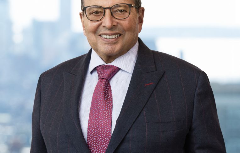 A smiling man in a suit in front of a window with buildings behind him.