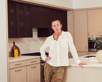 A man standing in a kitchen with one hand on the counter and another on his hip.
