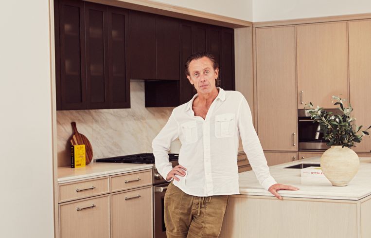A man standing in a kitchen with one hand on the counter and another on his hip.