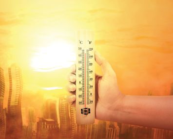 Hand holding thermometer with high temperature on the city with glowing sun background.