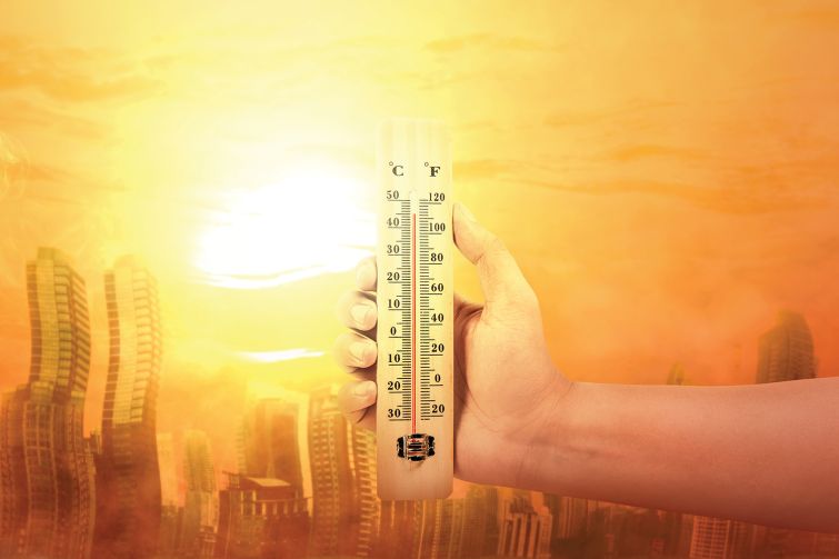 Hand holding thermometer with high temperature on the city with glowing sun background.