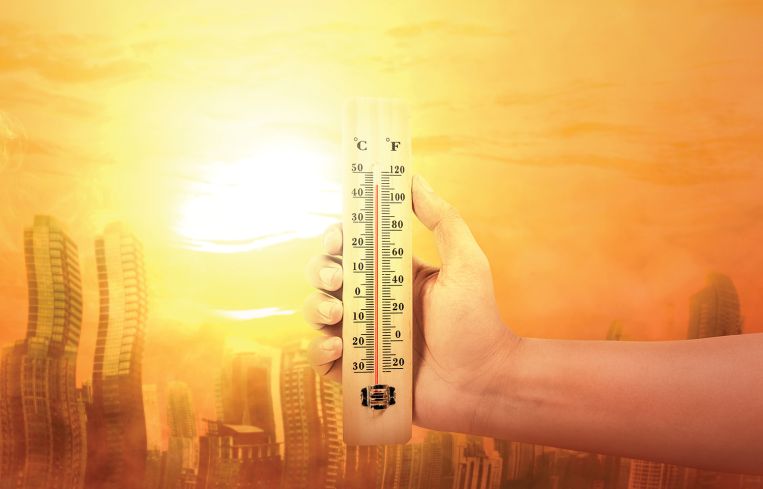 Hand holding thermometer with high temperature on the city with glowing sun background.