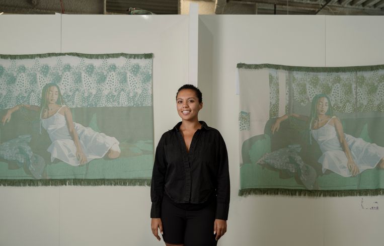 Malaika Temba stands in from of two of her textile pieces wearing a black hoodie.
