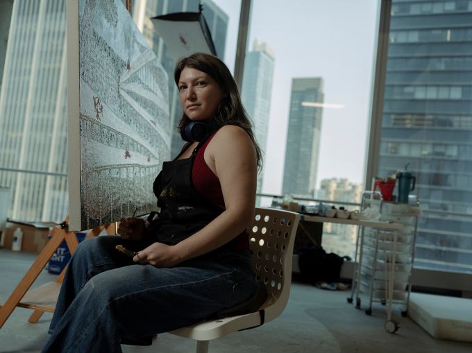 Anastasiya Tarasenko sits on a chair in front of a large canvas in front of a row of windows looking out onto Manhattan's Financial District.