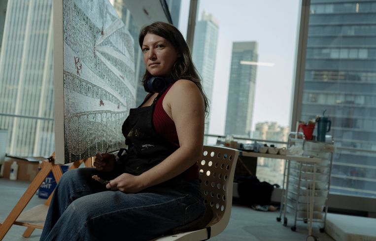 Anastasiya Tarasenko sits on a chair in front of a large canvas in front of a row of windows looking out onto Manhattan's Financial District.