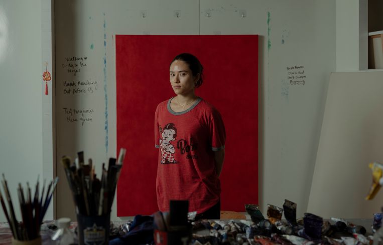 Gwen Hollingsworth stands in front of one of her oil paintings in a red T-shirt.