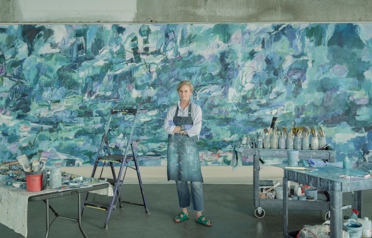 Kerry Irvine stands in front of an oversized canvas wearing a blue apron.