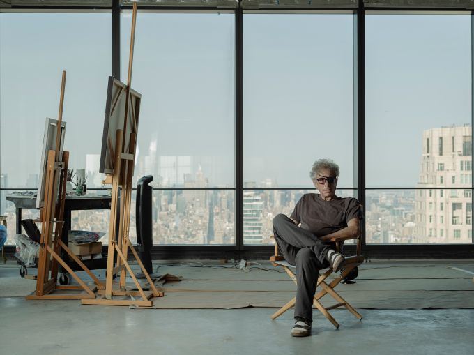 Todd Stone sits in a folding chair in front of a row of windows looking out onto the New York City skyline.