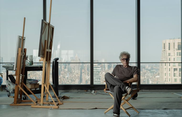 Todd Stone sits in a folding chair in front of a row of windows looking out onto the New York City skyline.