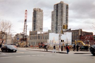 A construction site from a distance, with two towers rising behind it and a fence around it.