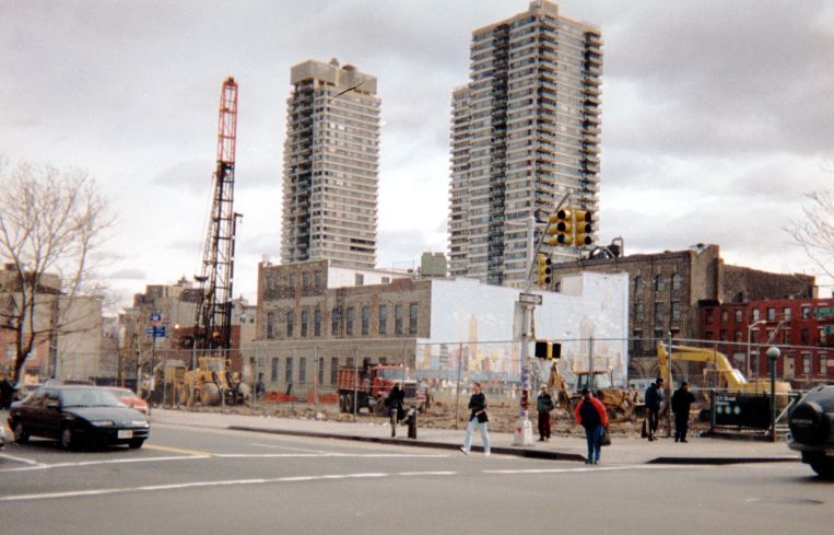 A construction site from a distance, with two towers rising behind it and a fence around it.