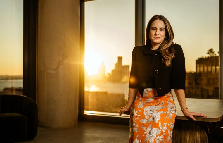 A woman standing at a desk with the sun setting behind her outside a building window.