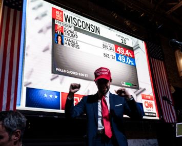 A man in a baseball cap celebrating election results on the screen behind him.