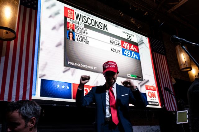 A man in a baseball cap celebrating election results on the screen behind him.