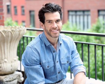 A man in an open-collar shirt on a terrace, smiling into the camera.