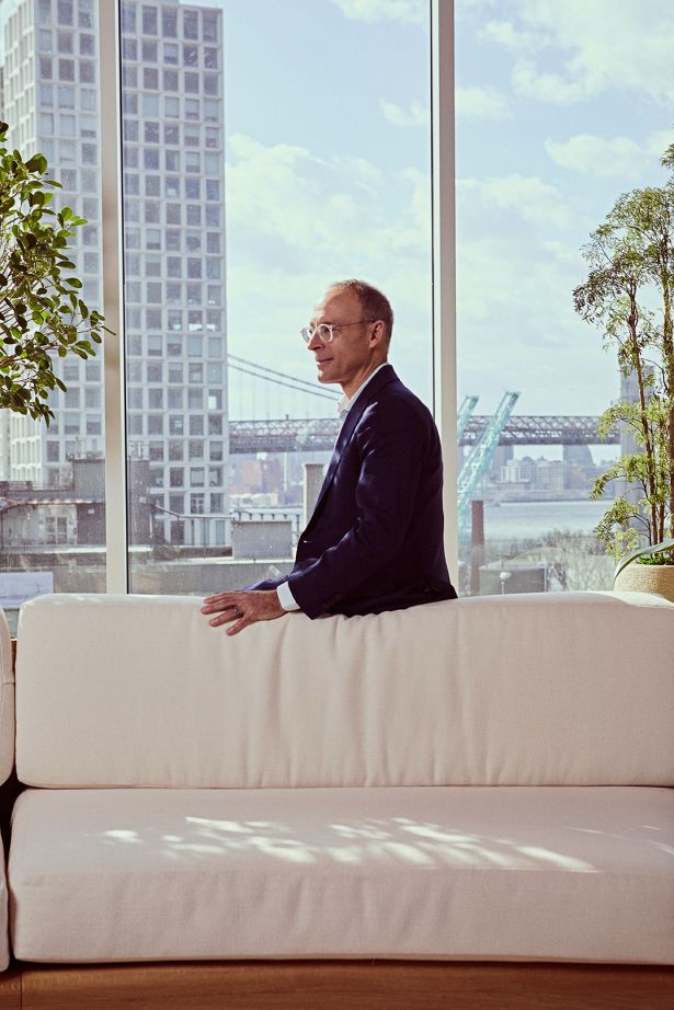 A man standing behind a couch with a building visible out the window behind him.