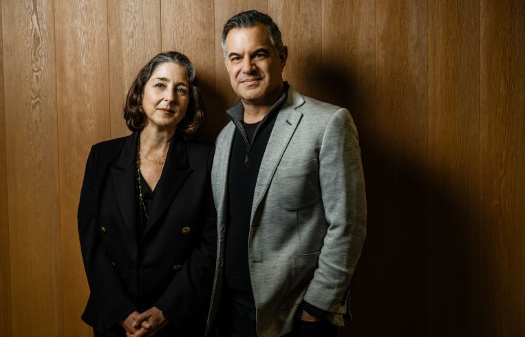 A woman and a man standing side by side in front of a wooden wall.