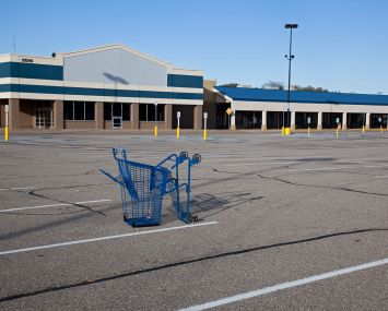 A shopping cart lies in a parking lot at a mostly-vacant shopping center.