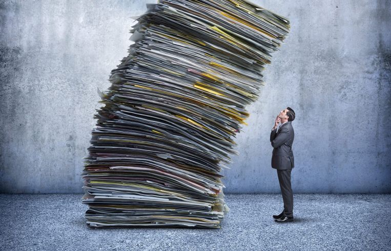 An illustration of a businessman looks up as a large stack of paperwork is leaning towards him.