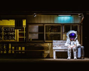 A person in a space suit sitting at a train station with a suitcase.