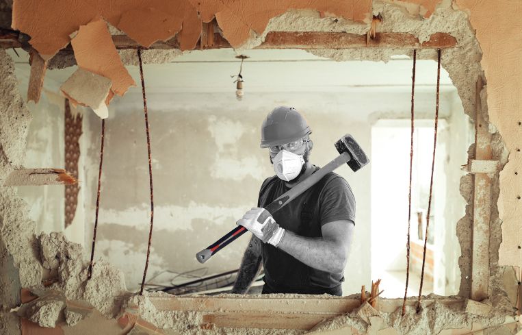 Builder with a hammer in his hands breaks the cement wall The builder is dressed in a protective suit and helmet.