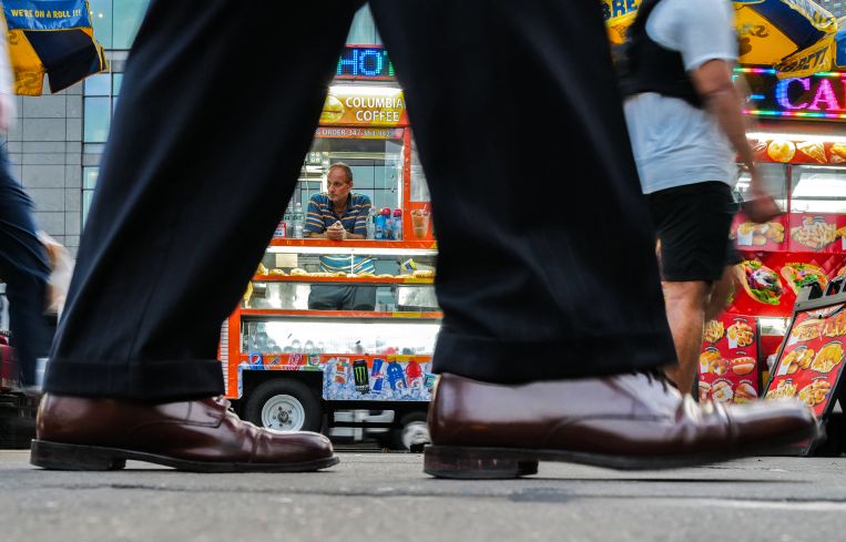 A hot dog and snack vendor in Manhattan waits for customers while pedestrians walk by.