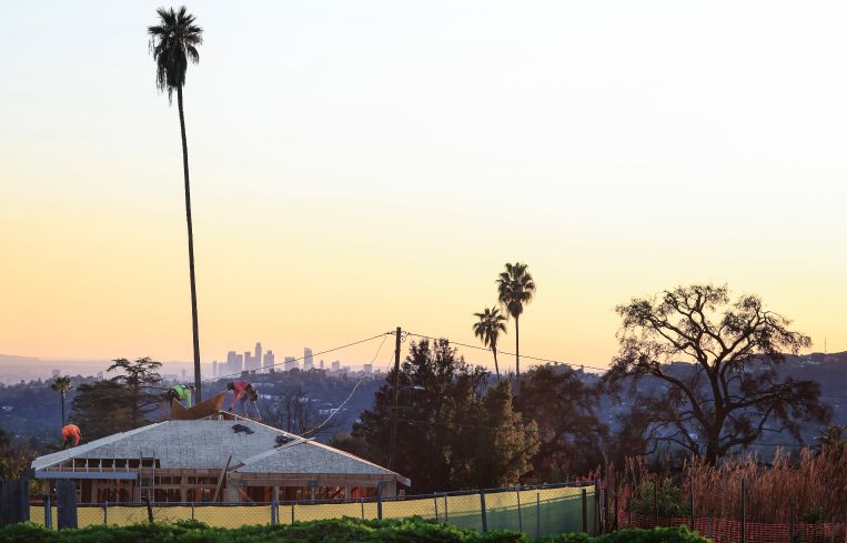 Workers in December help rebuild a home on a property destroyed in the Eaton Fire, with the Los Angeles downtown skyline visible beyond.