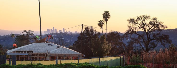 Workers in December help rebuild a home on a property destroyed in the Eaton Fire, with the Los Angeles downtown skyline visible beyond.