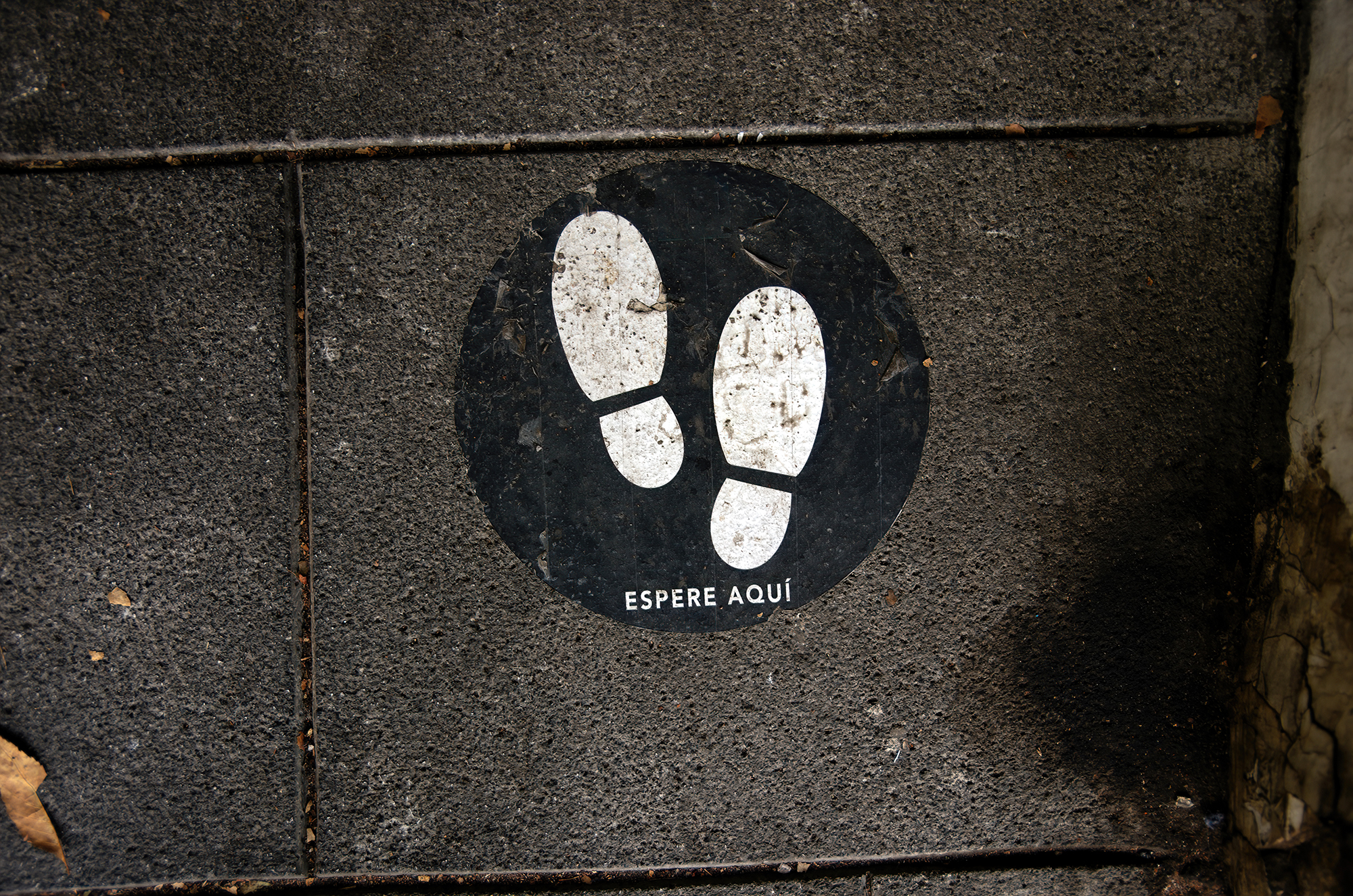 Spanish-language social distancing safety sticker on a concrete footpath stating 'Espere aquí' [Wait here]
