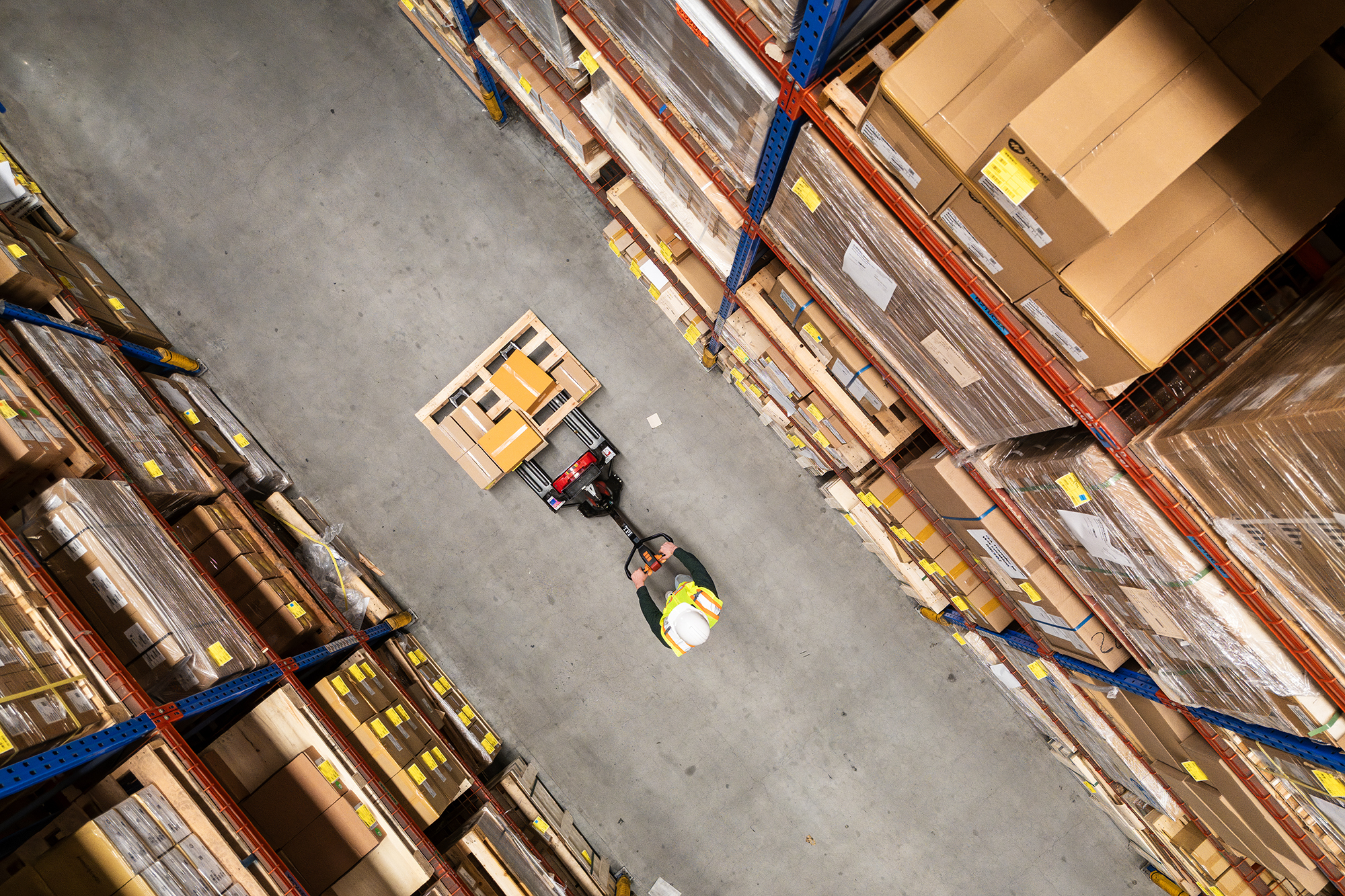 A warehouse worker navigates a forklift through rows of towering shelves filled with boxes.