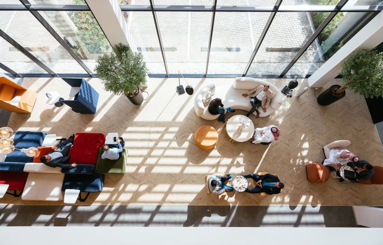Aerial view of a modern office lounge area with diverse people interacting and natural light streaming through large windows