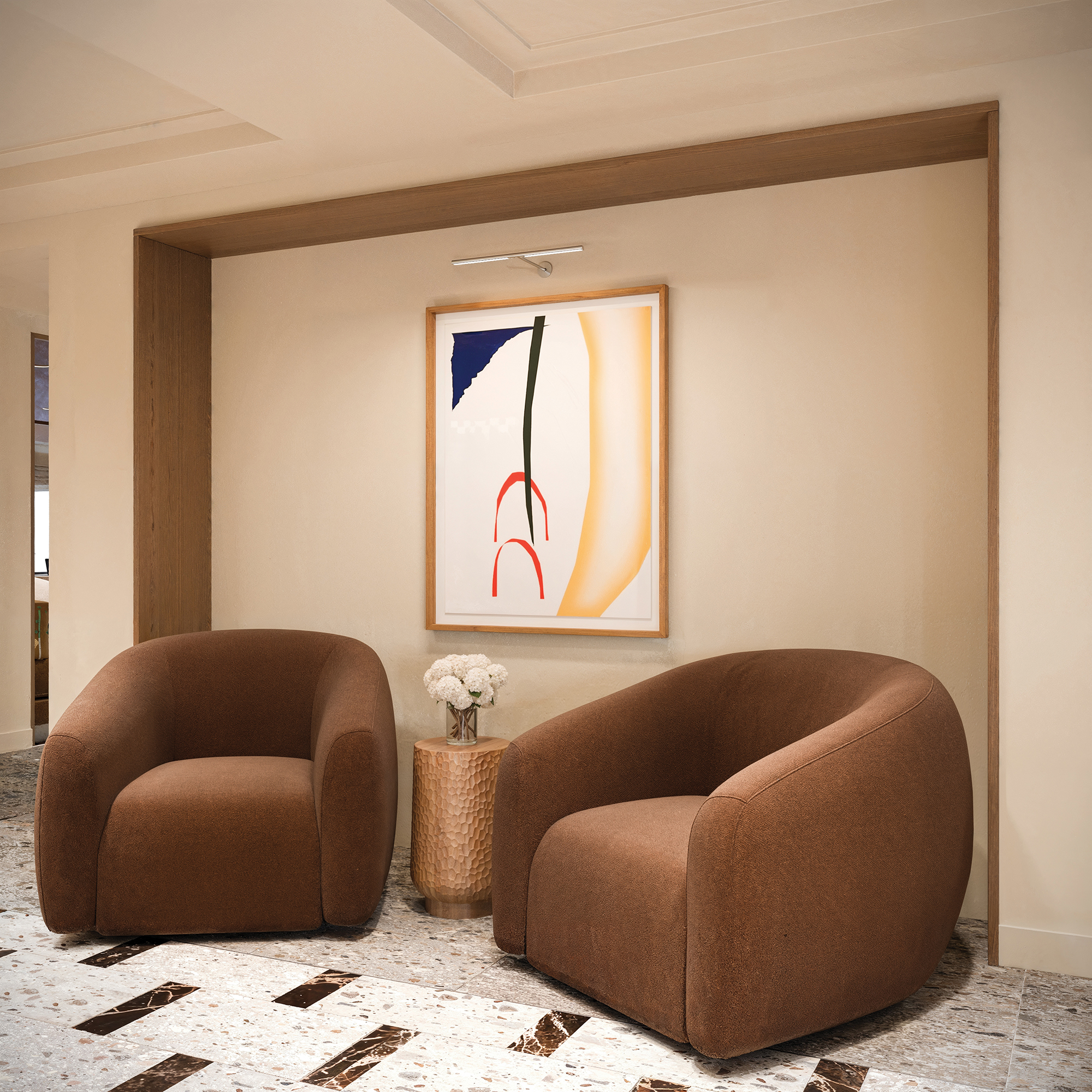 A pair of brown, round cushioned chairs sit in front of a beige wall with a minimalist artwork.
