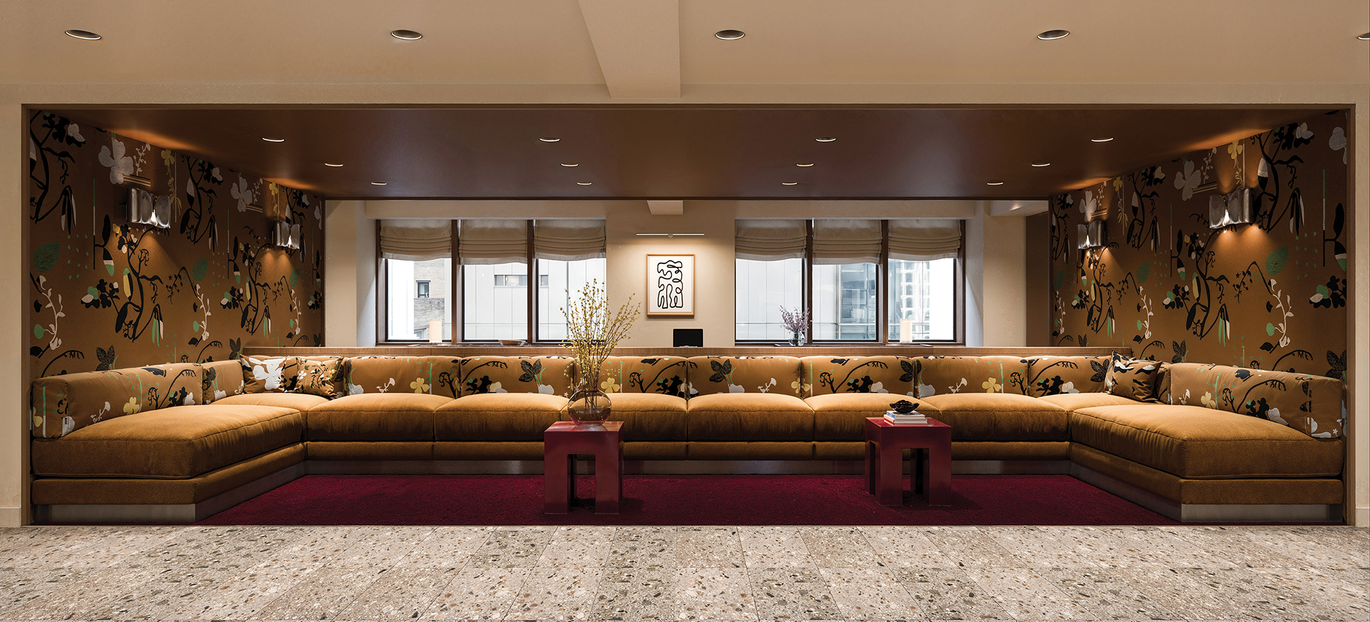An alcove features a line of windows, a burgundy carpet and a long, golden broad couch with floral cushions that match the wallpaper.