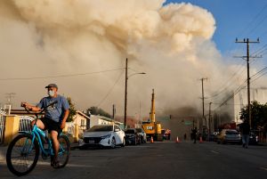 A person rides a bicycle as a massive warehouse fire at a Lineage cold storage facility burns on June 20 in Los Angeles.