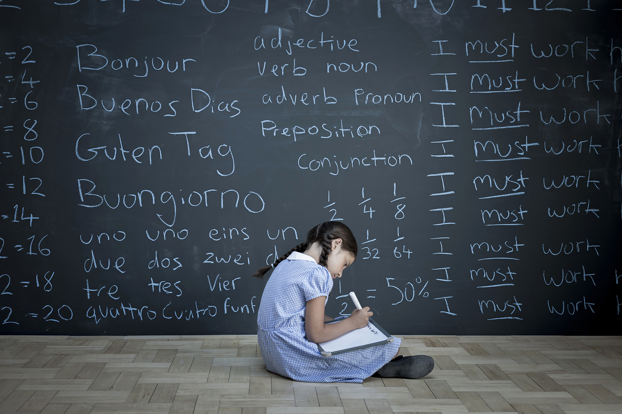 Schoolgirl sitting studying in front of large chalkboard with words and languages on it.